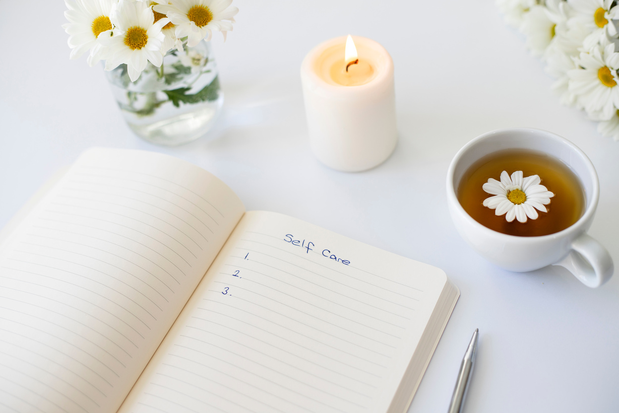 Close up of handwritten text "Self Care" in foreground with notebook, pen, cup of tea, flowers